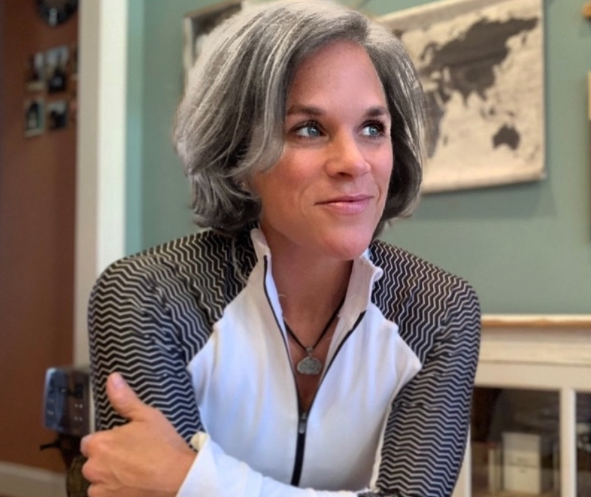 Woman with short gray hair, wearing a white and black patterned jacket, sitting indoors with a world map in the background.