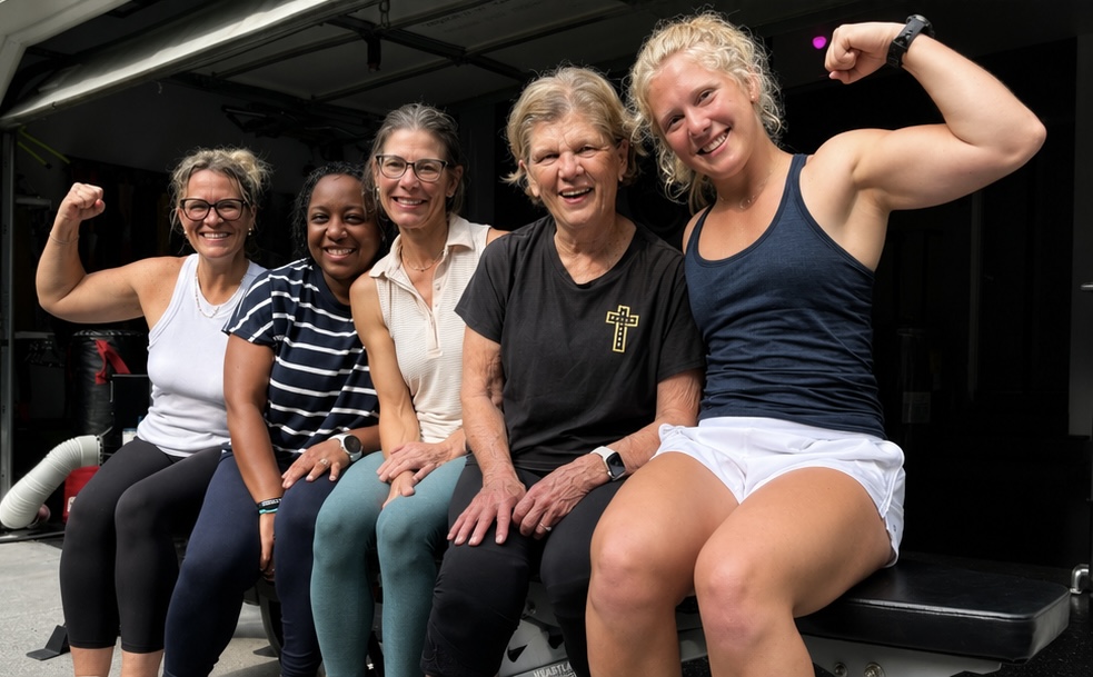 Five women sitting together, smiling, with two women flexing their muscles, in a casual setting.