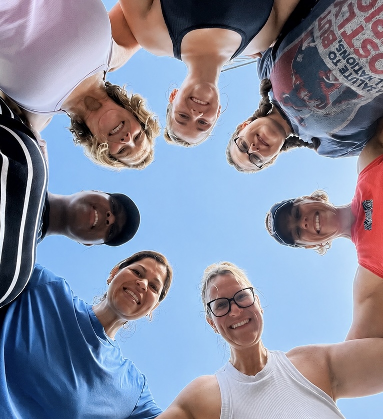 Group of people standing in a circle, looking down at the camera, smiling, against a blue sky.