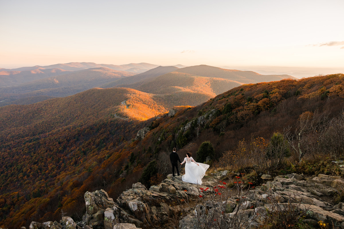 Person in a white dress standing on rocky mountain edge with a landscape of rolling hills and mountains in the background.