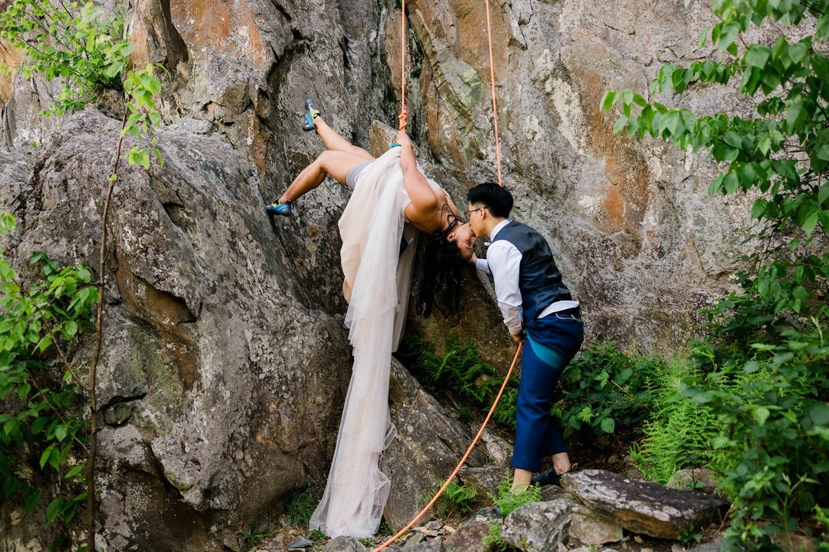 A person in a white dress is hanging upside down on a rocky cliff, leaning toward another person in a vest and pants, holding a rope, surrounded by greenery.