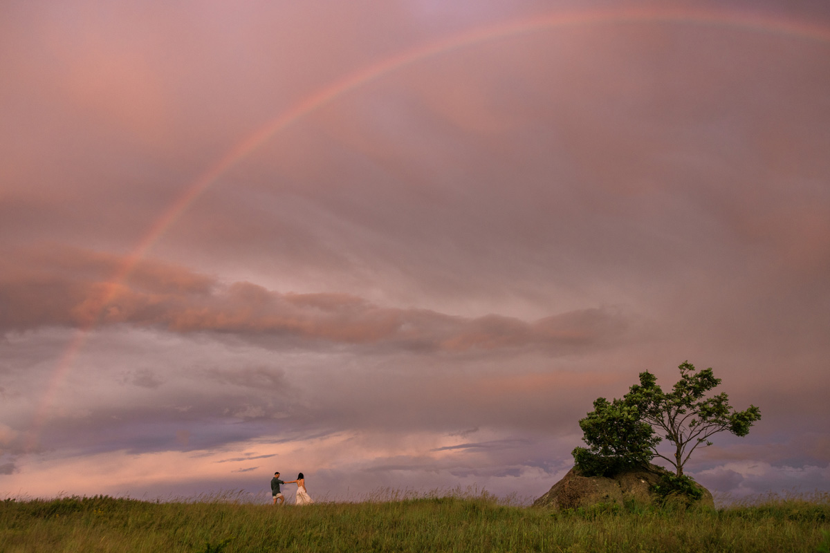 Two people standing on a grassy field with a rainbow in the sky and a tree on the right.