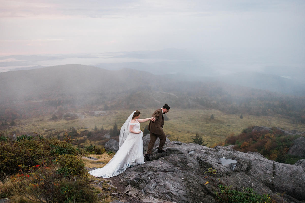 A bride and groom walk on a rocky hilltop with a misty landscape in the background, holding hands and looking at each other.