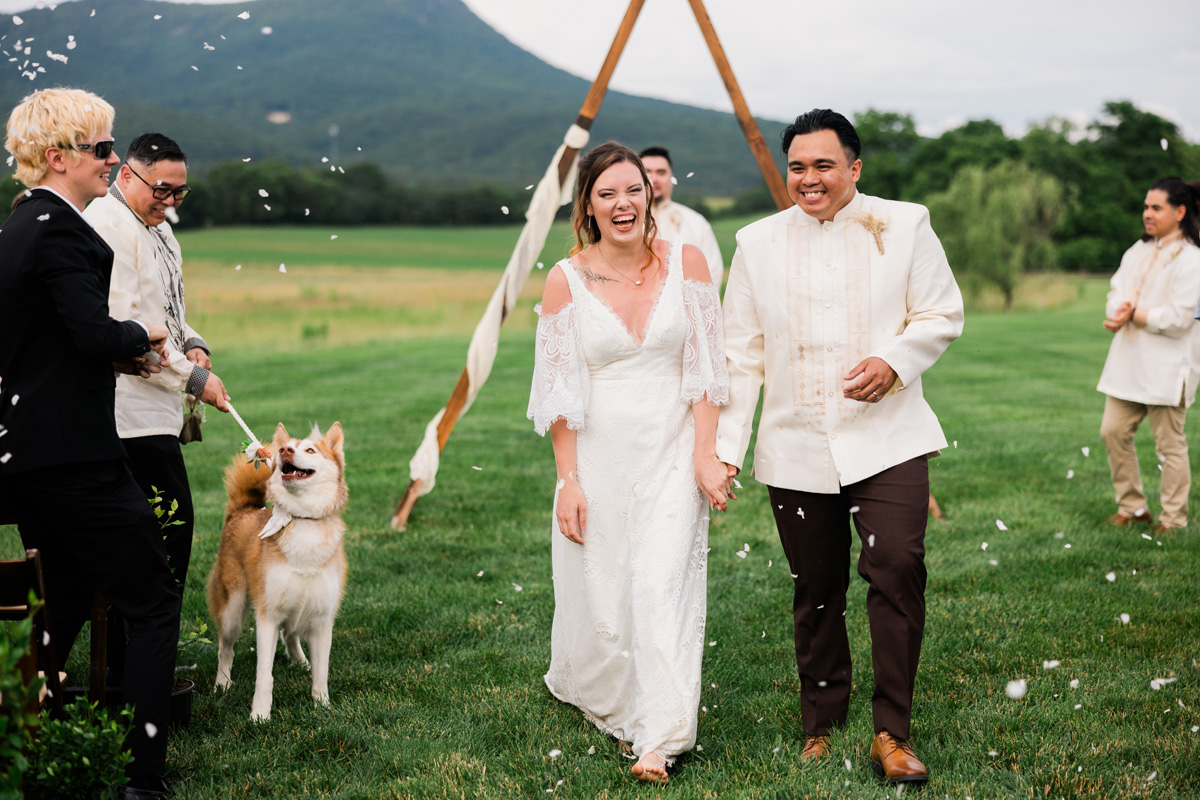 Couple in wedding attire walking outdoors with friends, a dog, and a scenic mountain background, celebrating a special occasion.