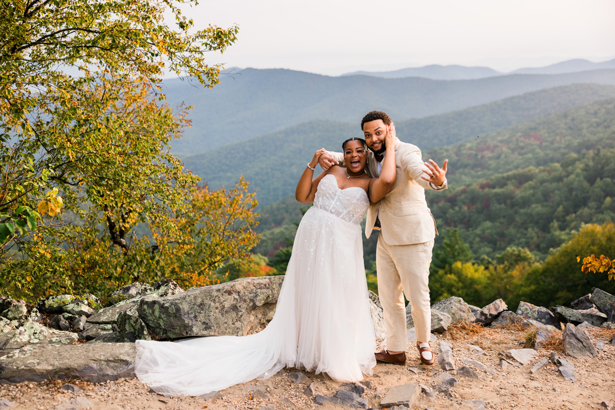 A newlywed couple stands on rocks outdoors with mountains and trees in the background, smiling and celebrating.
