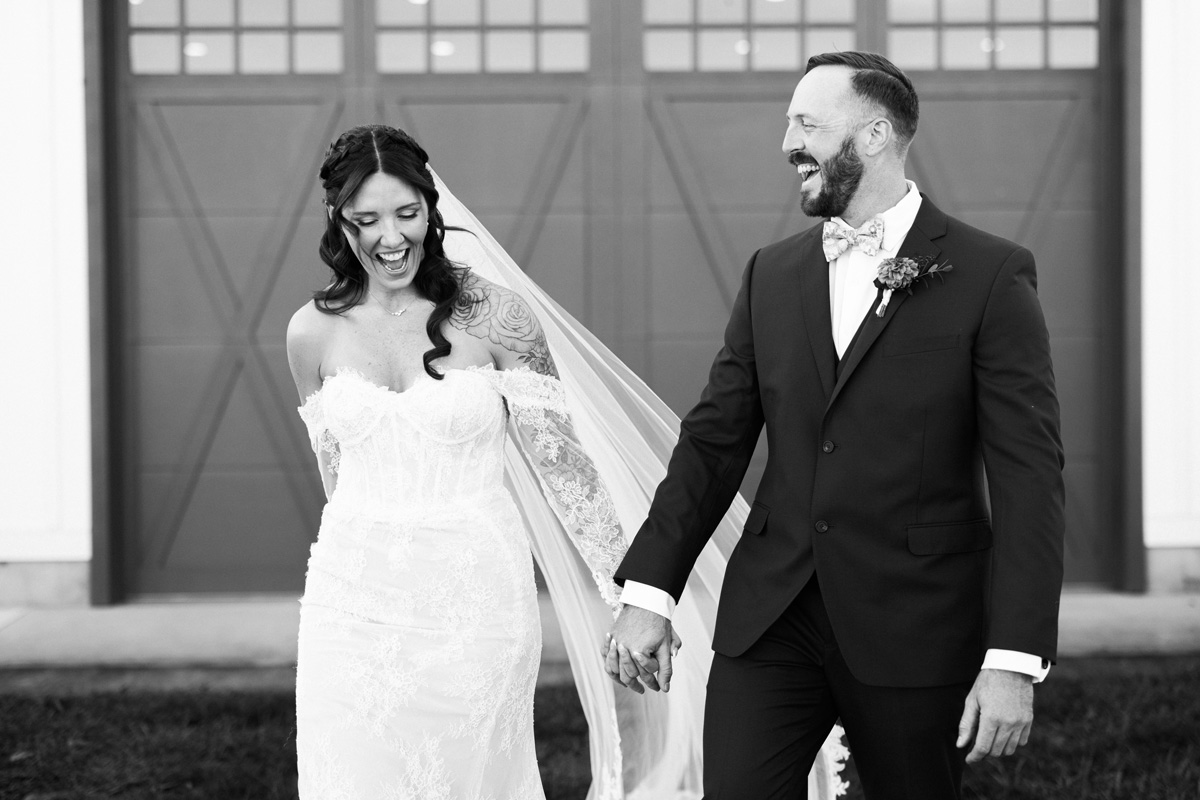 Bride and groom holding hands, smiling, standing outside in front of a large door, in black and white.