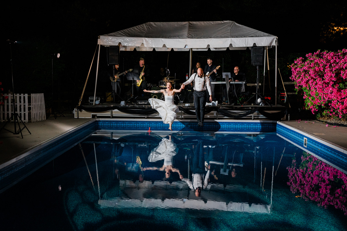 People dancing by a pool with a band playing on a stage in the background at night, reflected in the water.