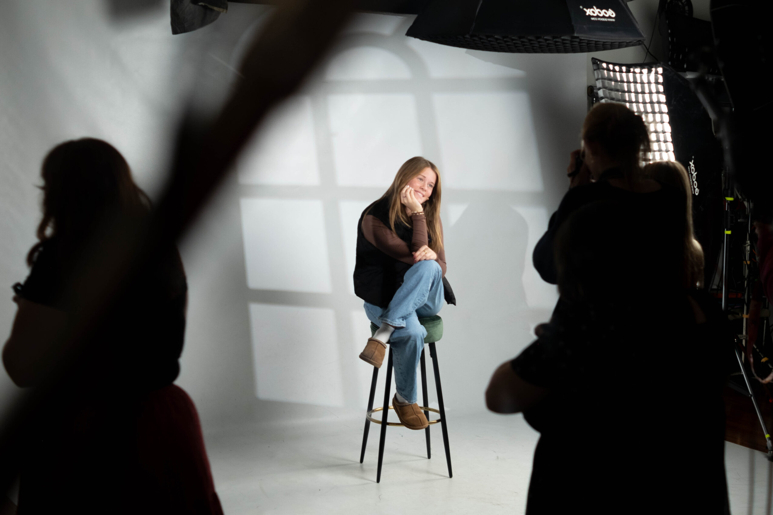 Person sitting on a stool in a photography studio with a white backdrop, surrounded by people and studio equipment.