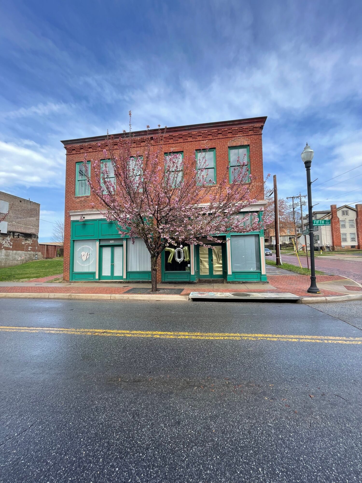 Three-story brick building with large windows, a tree with pink blossoms, and a green storefront on a street corner.