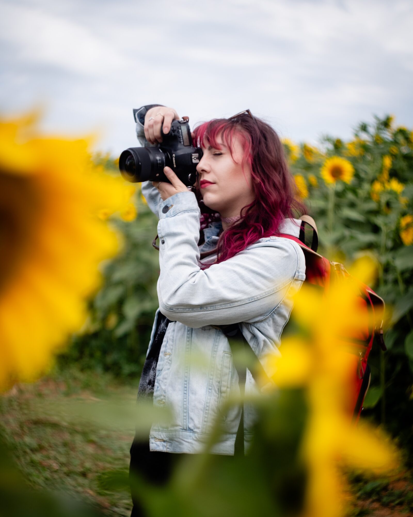 Young woman with red hair holding a camera in a sunflower field, looking through the viewfinder, cloudy sky background.