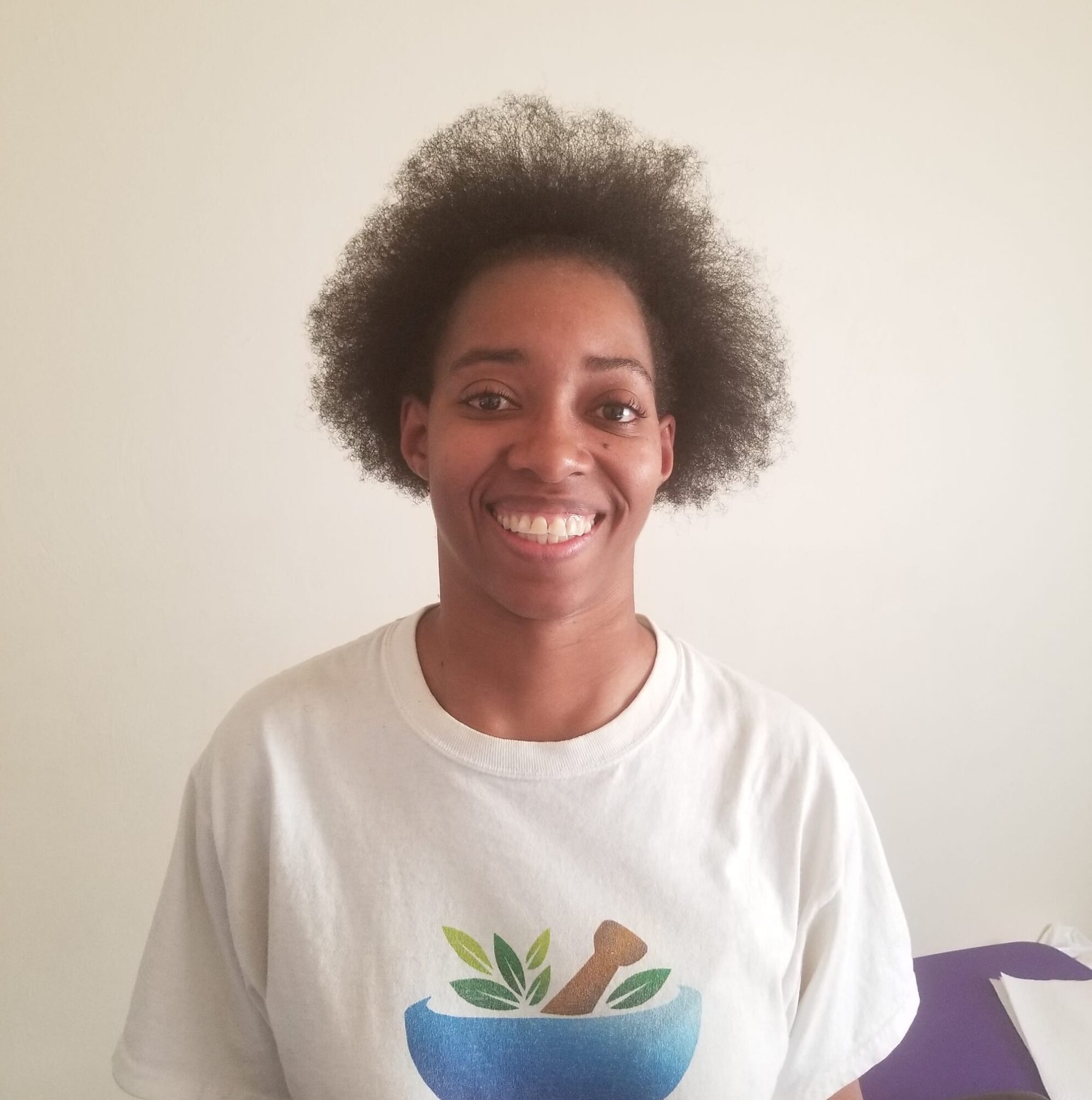 Smiling person with natural hair wearing a white T-shirt with a plant and bone illustration, standing against a plain wall.