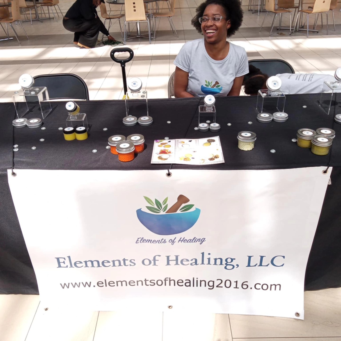 Smiling woman sitting at a table with jars and a brochure, representing Elements of Healing, LLC, at an indoor event.