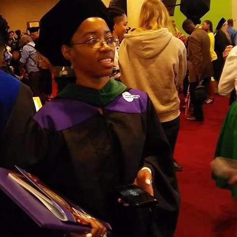Woman in graduation gown and cap holding a phone, surrounded by people at an indoor event.