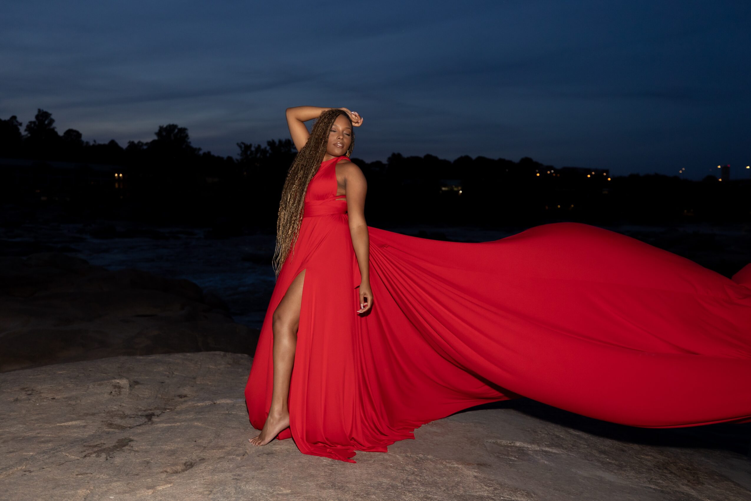 Woman in a red gown with a long train standing on a beach at dusk, holding her hair back, with water and trees in background.