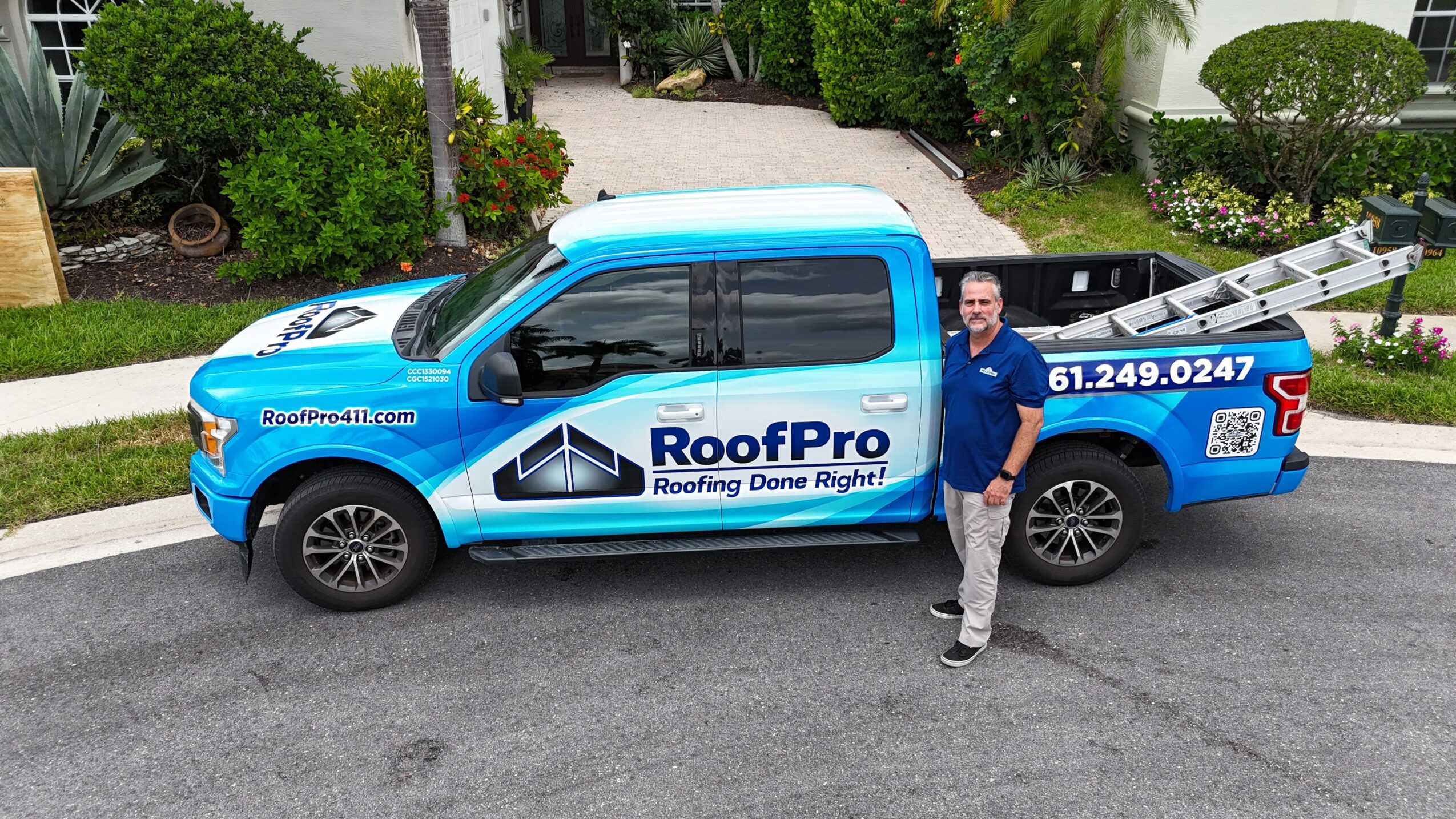 Man standing next to a blue roofing company truck on a residential street, with greenery and houses in background.