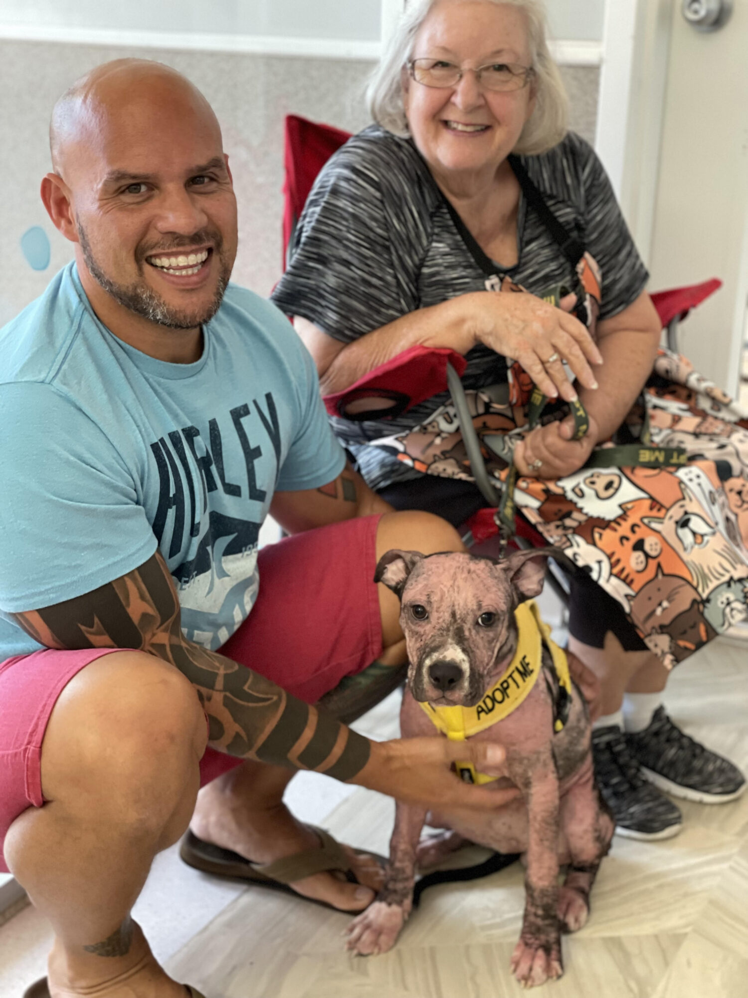 A smiling man, an elderly woman in a wheelchair, and a dog with a yellow vest sit together indoors.