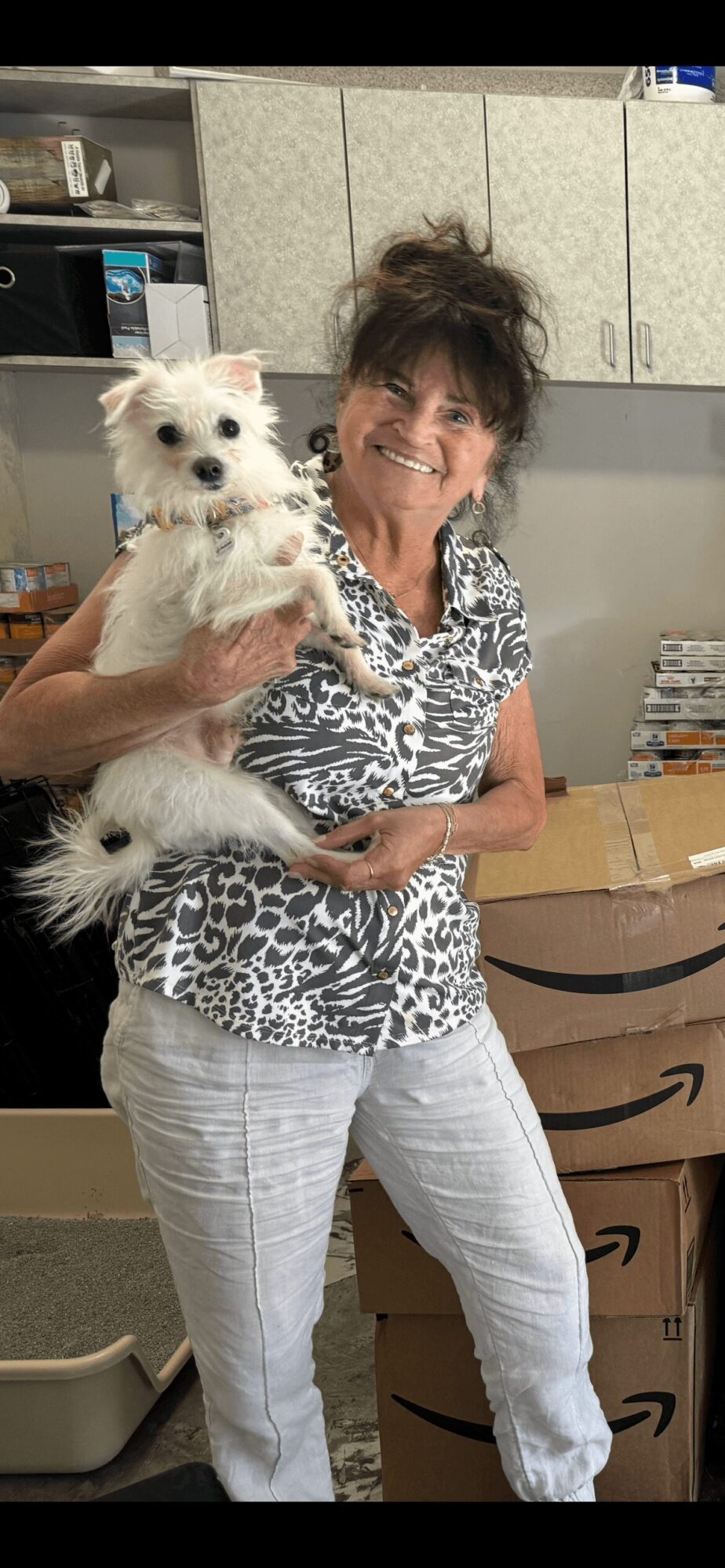 Woman holding a small white dog indoors, smiling, with cabinets and boxes in the background.