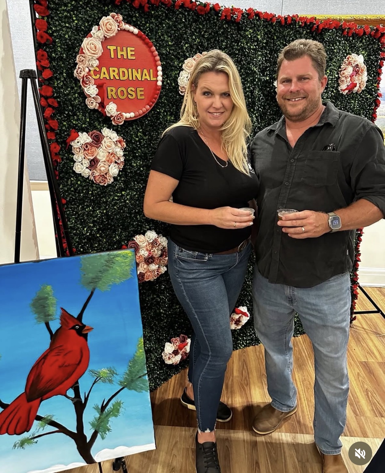 A woman and man stand together holding drinks in front of a floral backdrop with a sign reading 'The Cardinal Rose.'