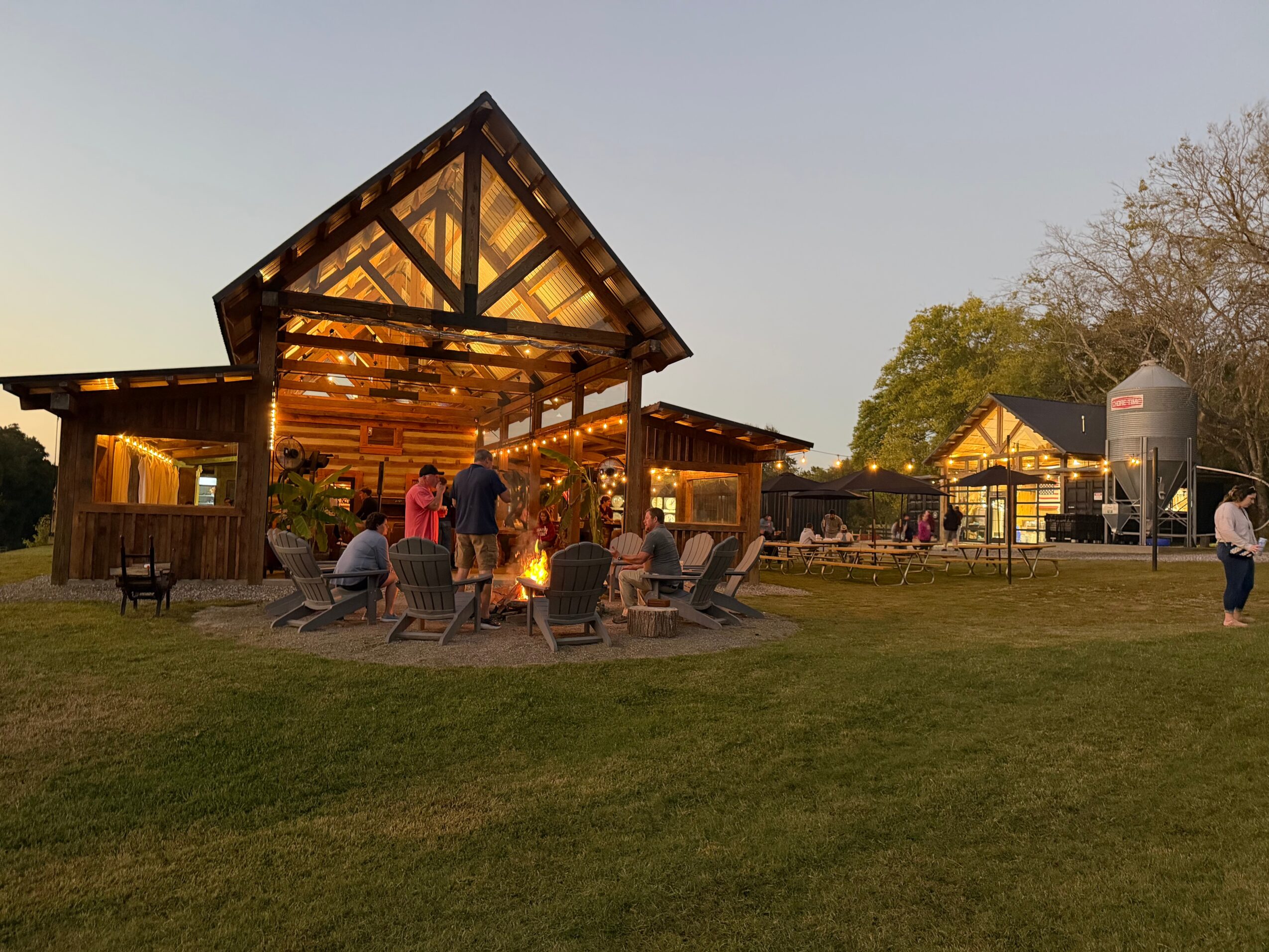 People gather around outdoor tables near a large wooden pavilion with a peaked roof, trees in the background, during evening light.