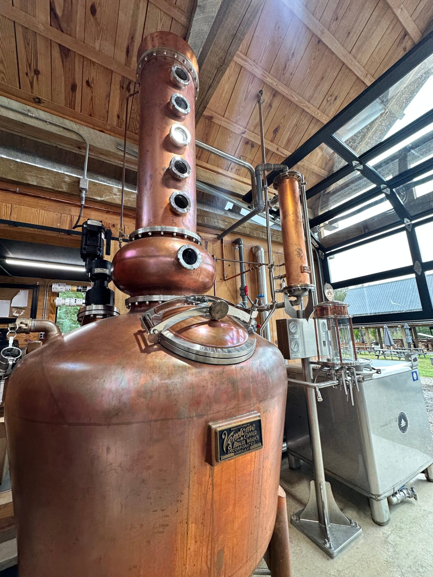 Copper still with a long, curved neck and a thermometer, situated in a wooden room with large windows.