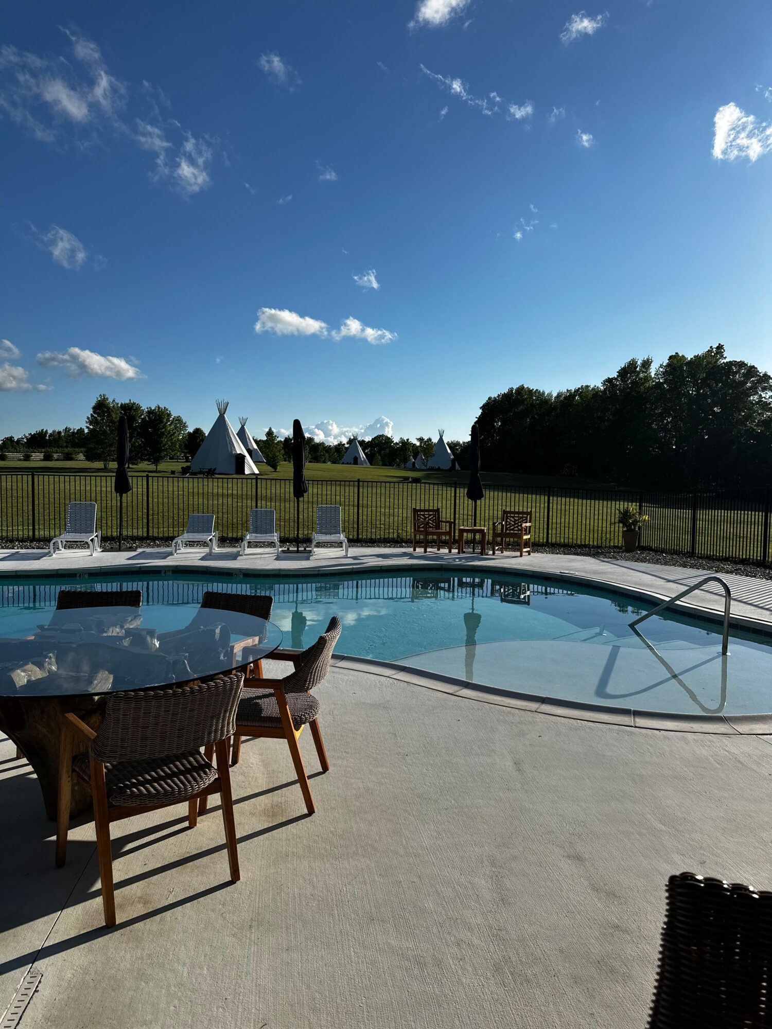Swimming pool with chairs, fence, trees, and blue sky with clouds, viewed from the side. Bright and sunny day.