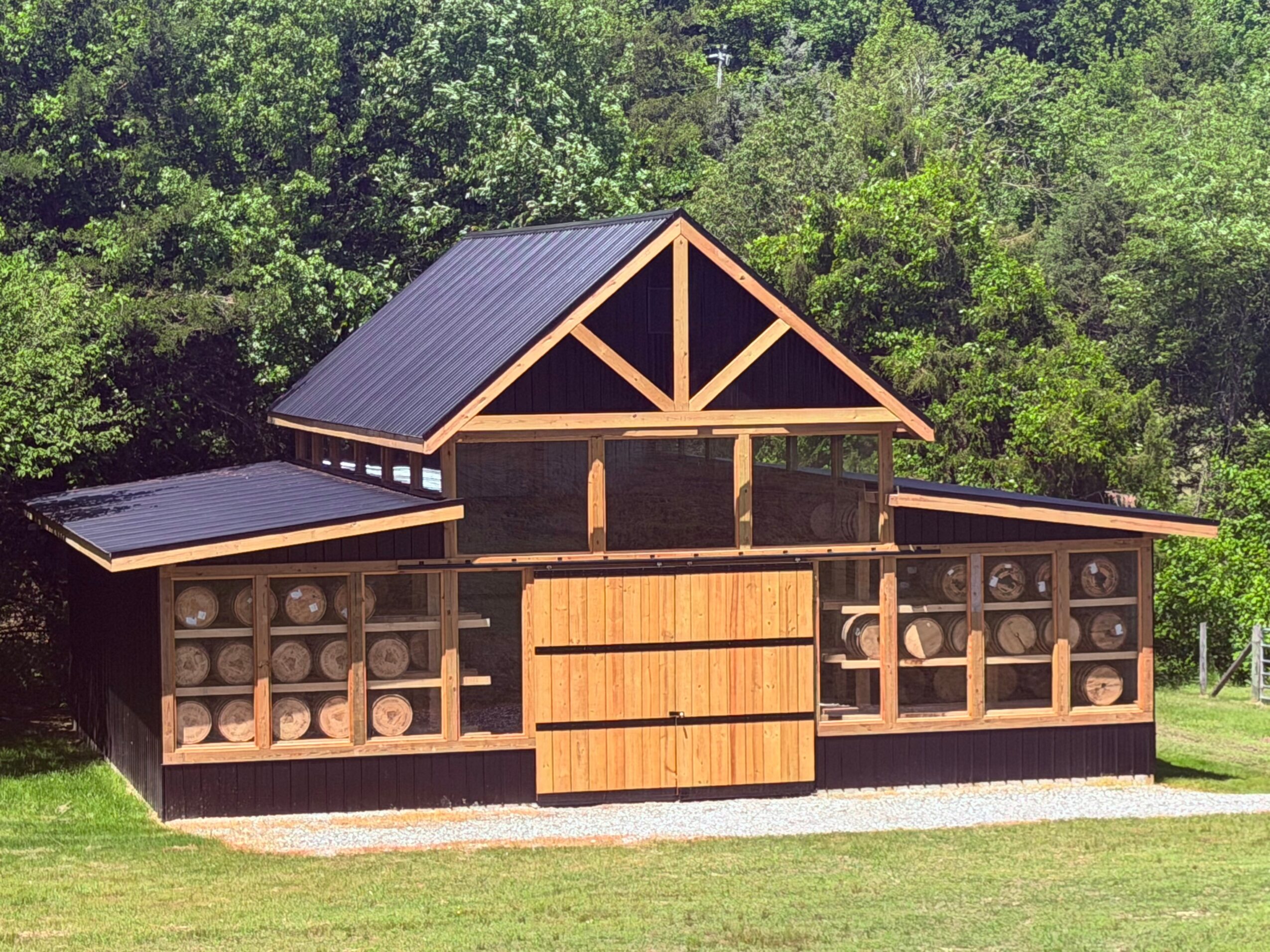 Wooden barn with a gabled roof, surrounded by green trees and grass, with a large sliding door in the center.