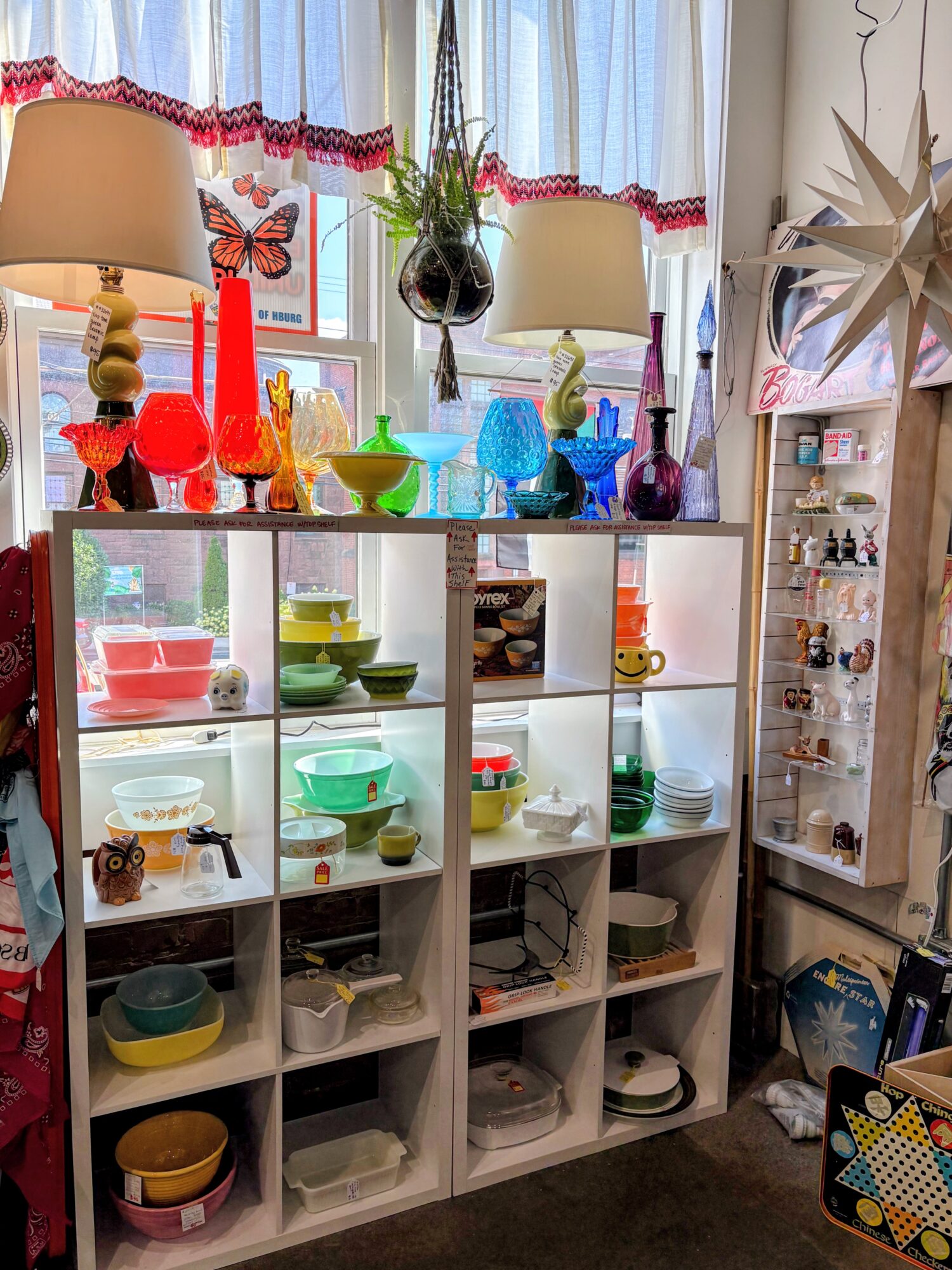 Display shelf with colorful glassware, bowls, and decorative items, near a window with curtains and a hanging plant.