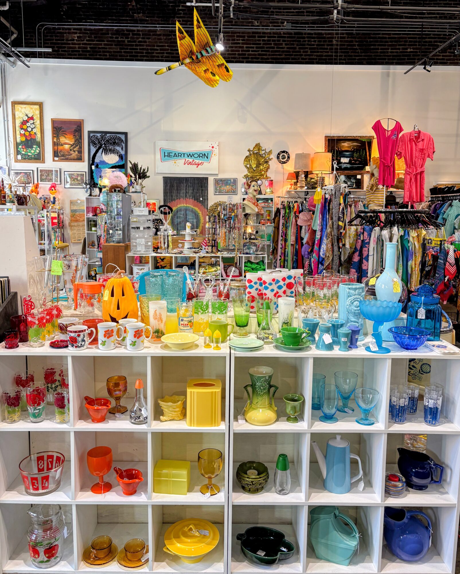 Store display with colorful glassware, ceramics, and decorative items on white shelves, clothing racks, and a yellow airplane hanging from the ceiling.