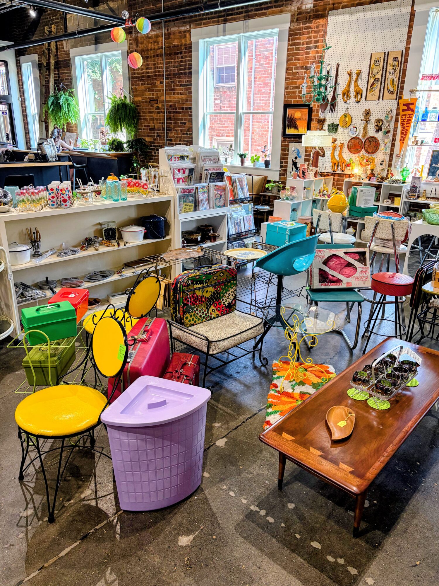 Interior of a store with colorful chairs, tables, and shelves filled with various items, large windows, and brick walls.
