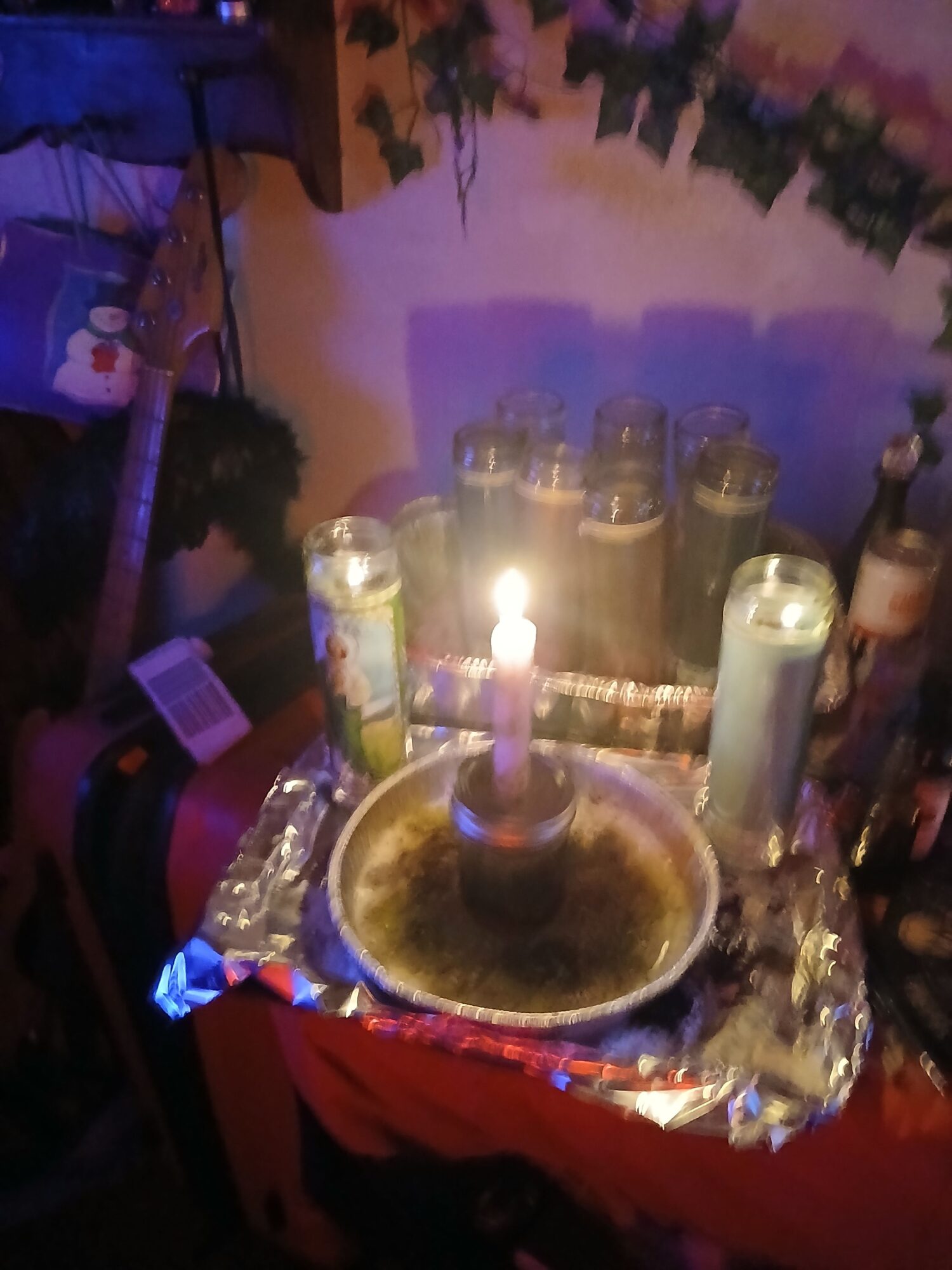 A table with bottles, a lit candle, and a bowl, set against a colorful background with plants.