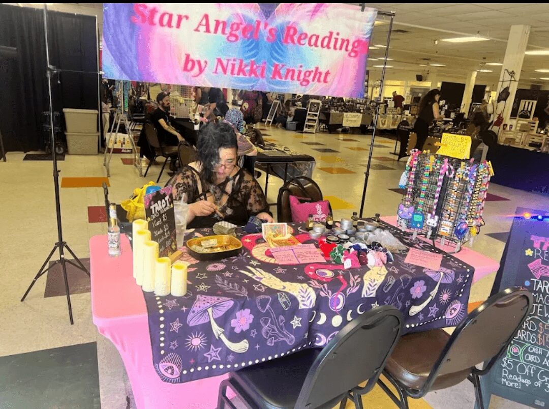 Person sitting at a table with candles, jewelry, and decorations, reading a book or sign, in a shopping mall setting.