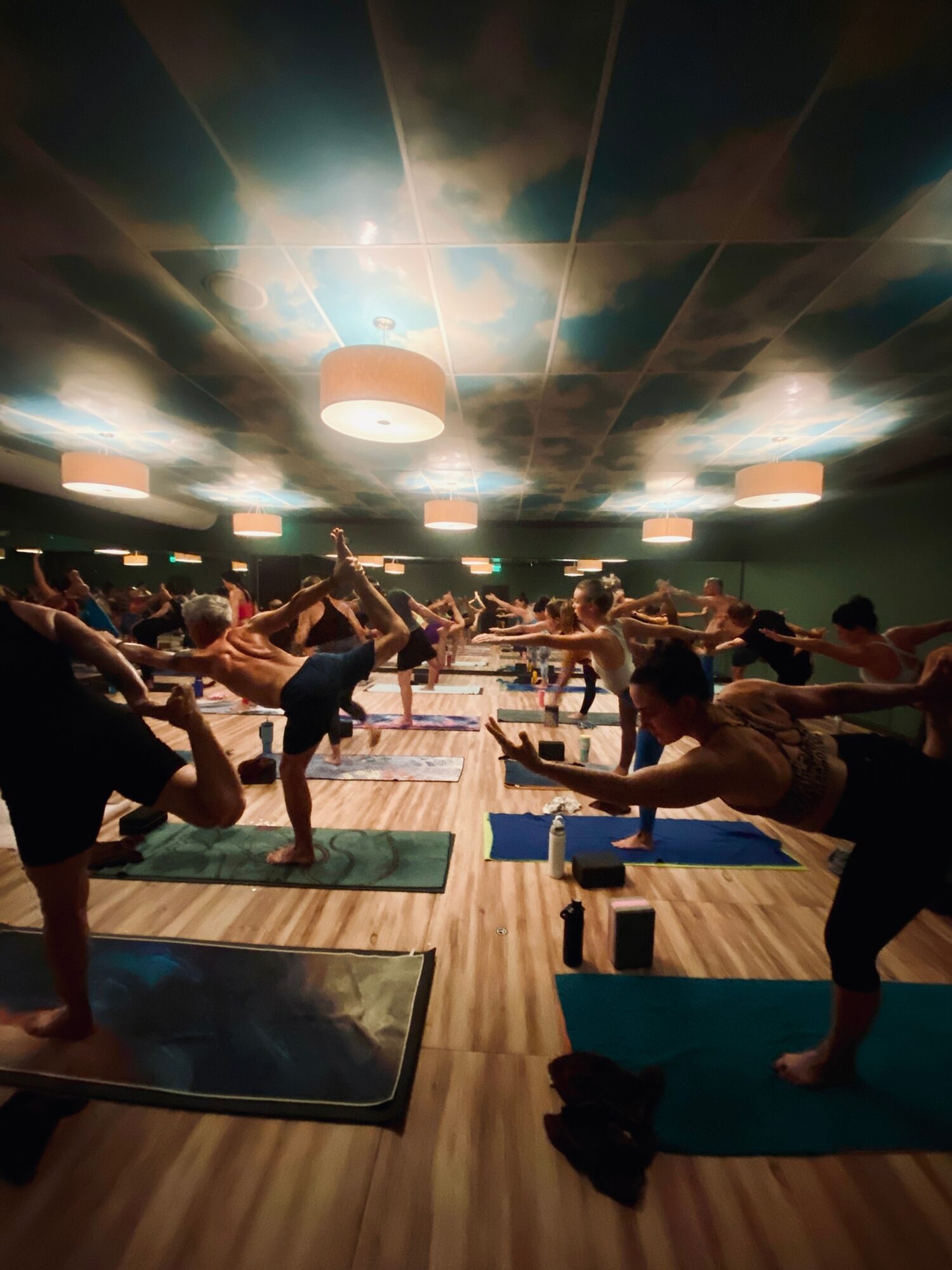 Group of people practicing yoga in a dimly lit studio with a reflective ceiling, mats on the floor, and participants stretching.