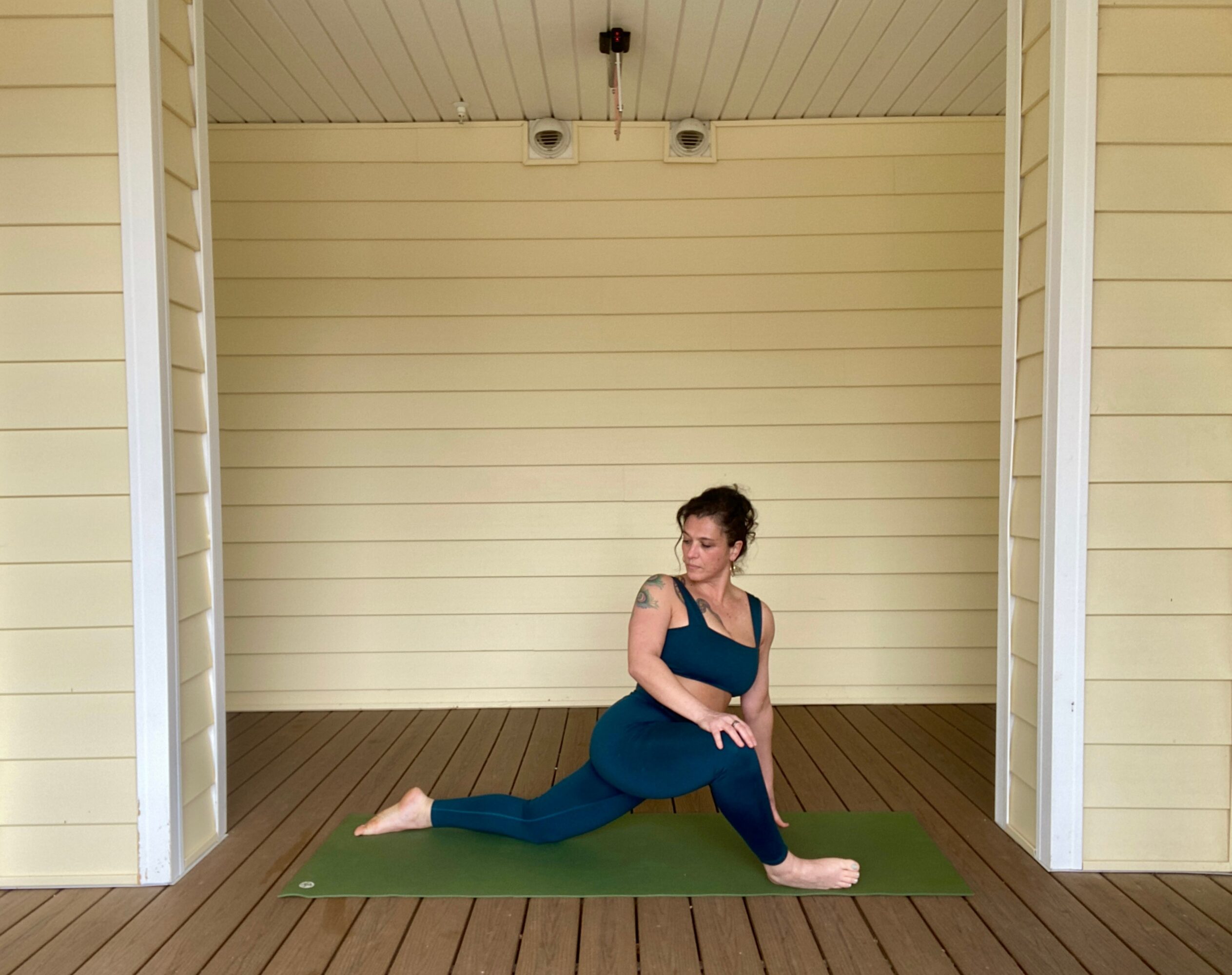 Woman practicing yoga on a green mat outdoors, sitting with one leg extended and the other bent, against a yellow wall.