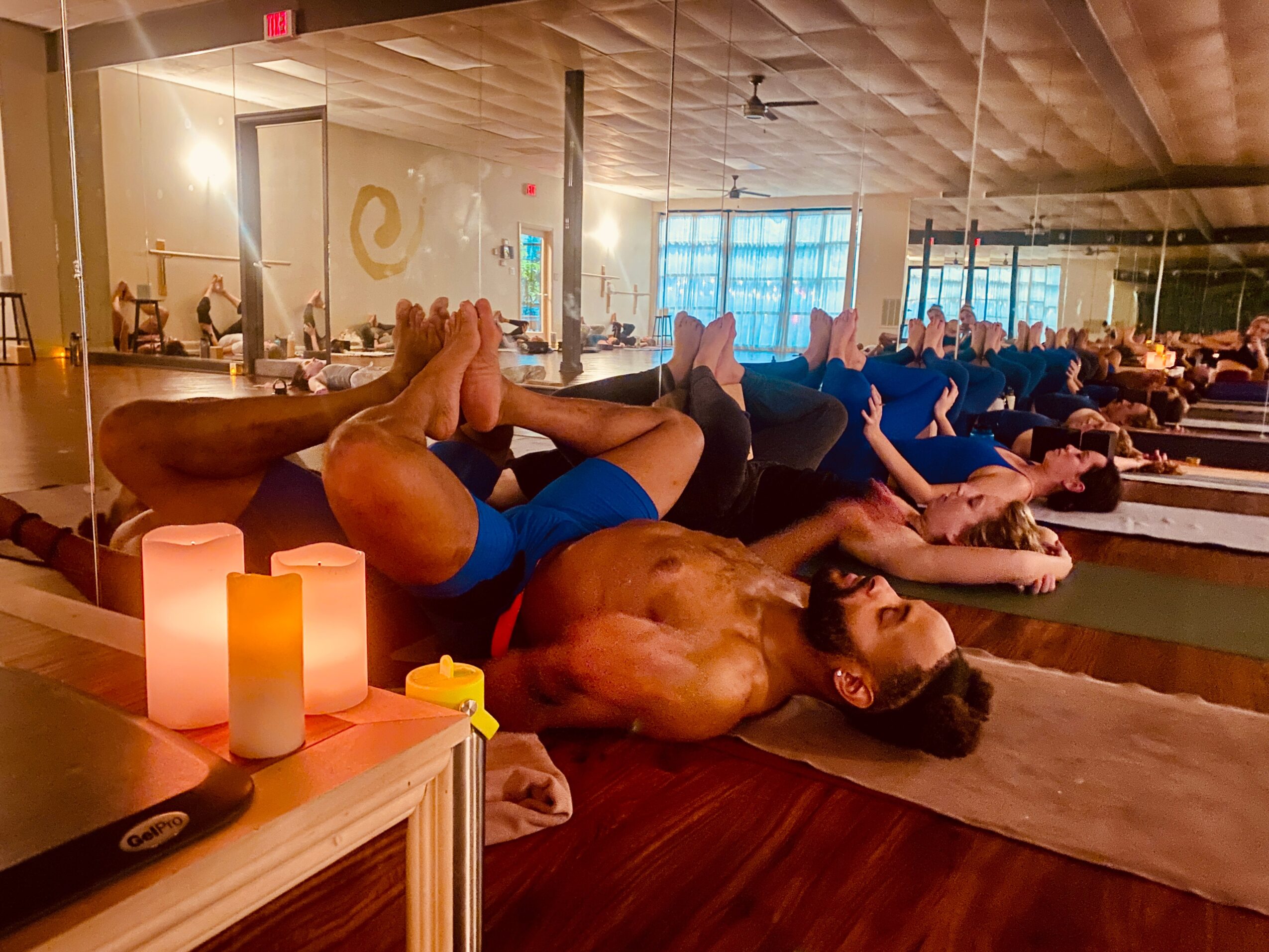 People practicing yoga on mats in a spacious, well-lit studio with large windows and wooden floors.