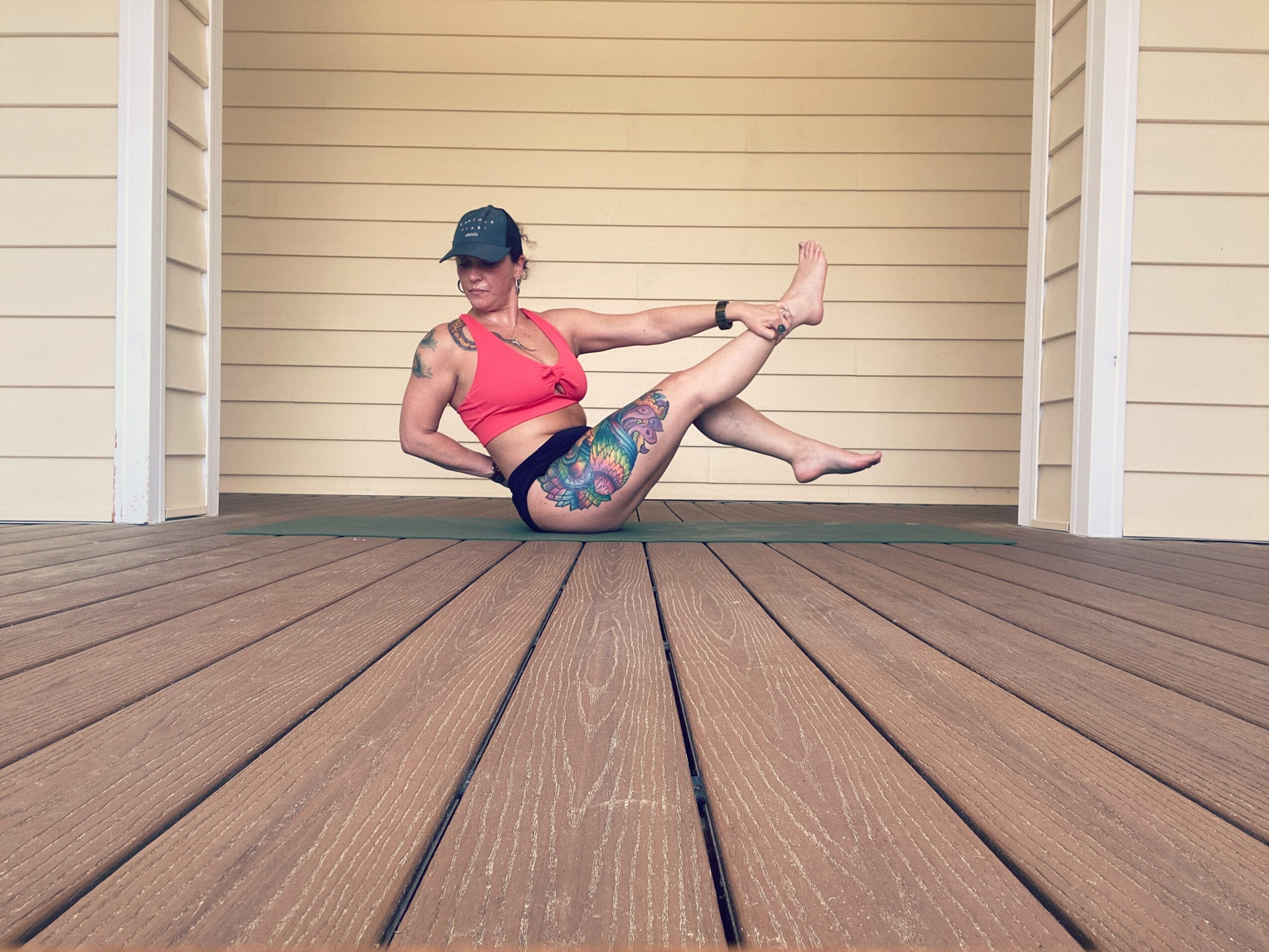 Person in a pink sports bra and colorful leggings sitting on a mat, leaning back with legs extended, on a wooden deck.