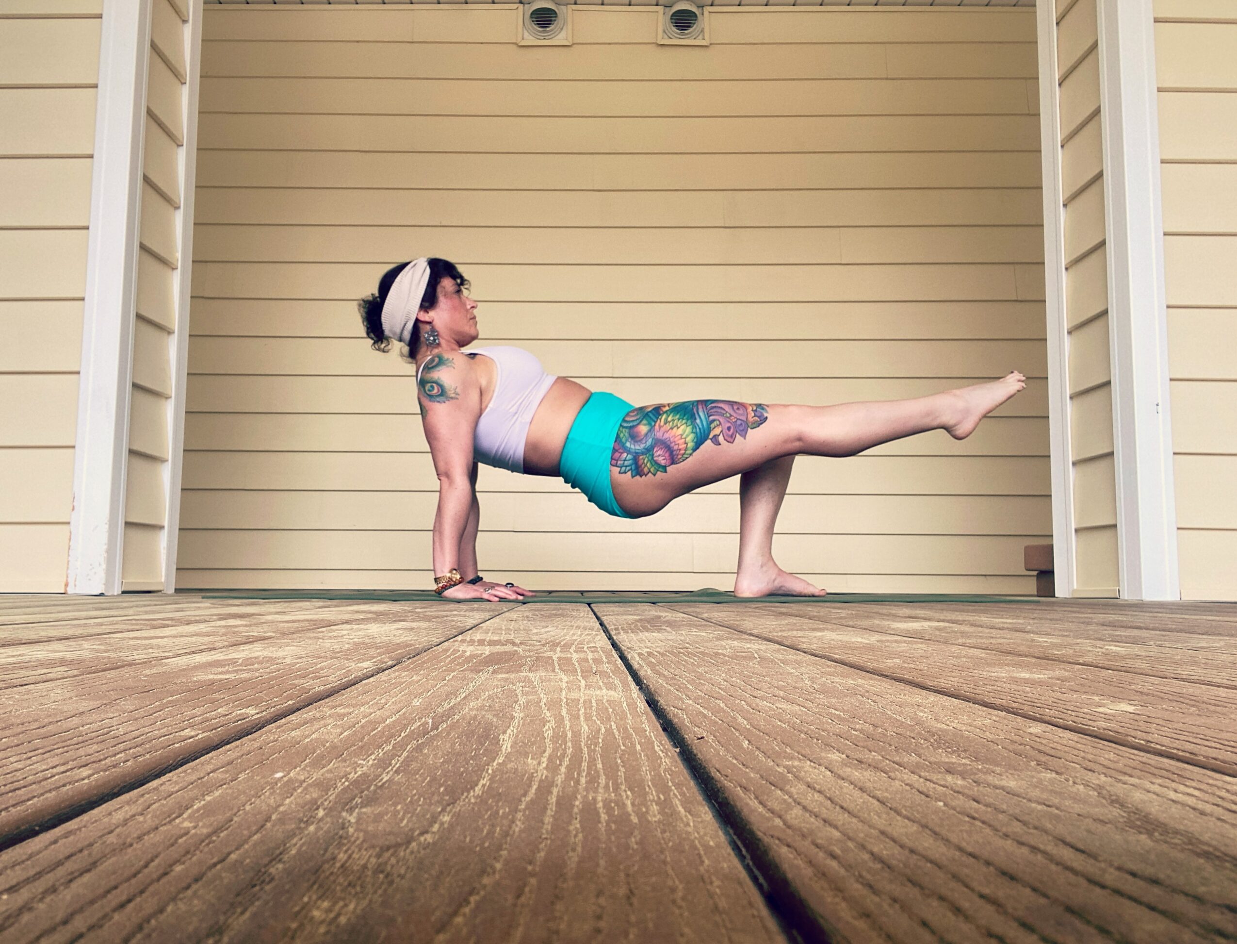 Person performing a yoga pose outdoors on wooden deck, with tattoos and wearing a headband, against a beige garage door.