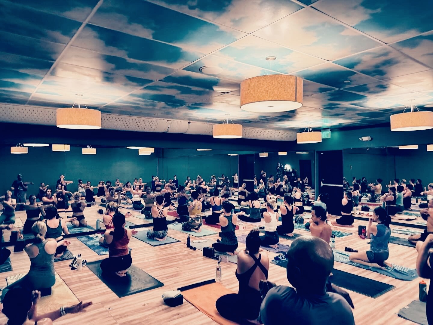 Large indoor yoga class with people practicing on mats, facing forward, in a spacious room with a sky-themed ceiling.
