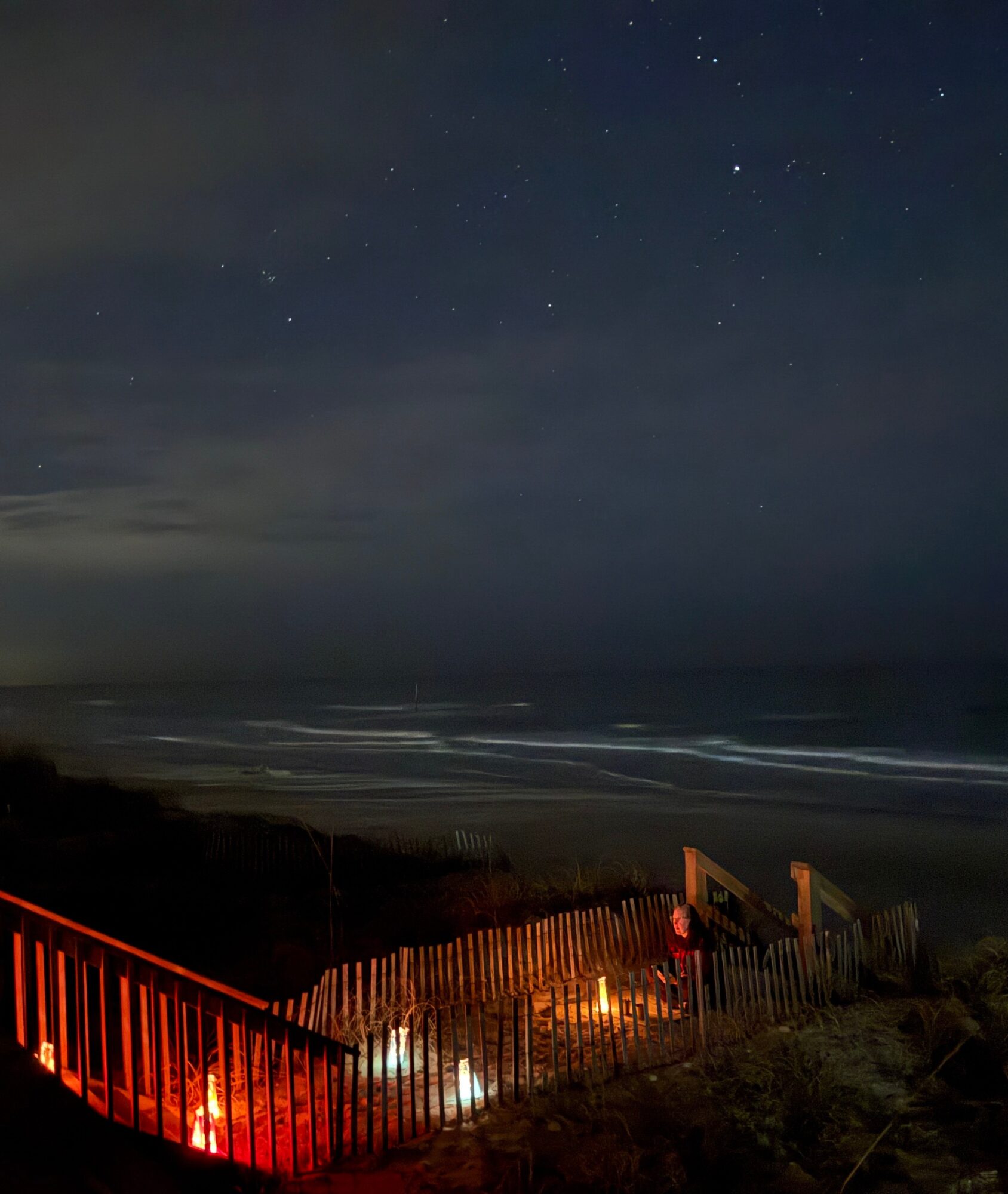 Nighttime beach scene with stars in the sky, ocean waves, and a fenced area with lights and a person, 20 words.