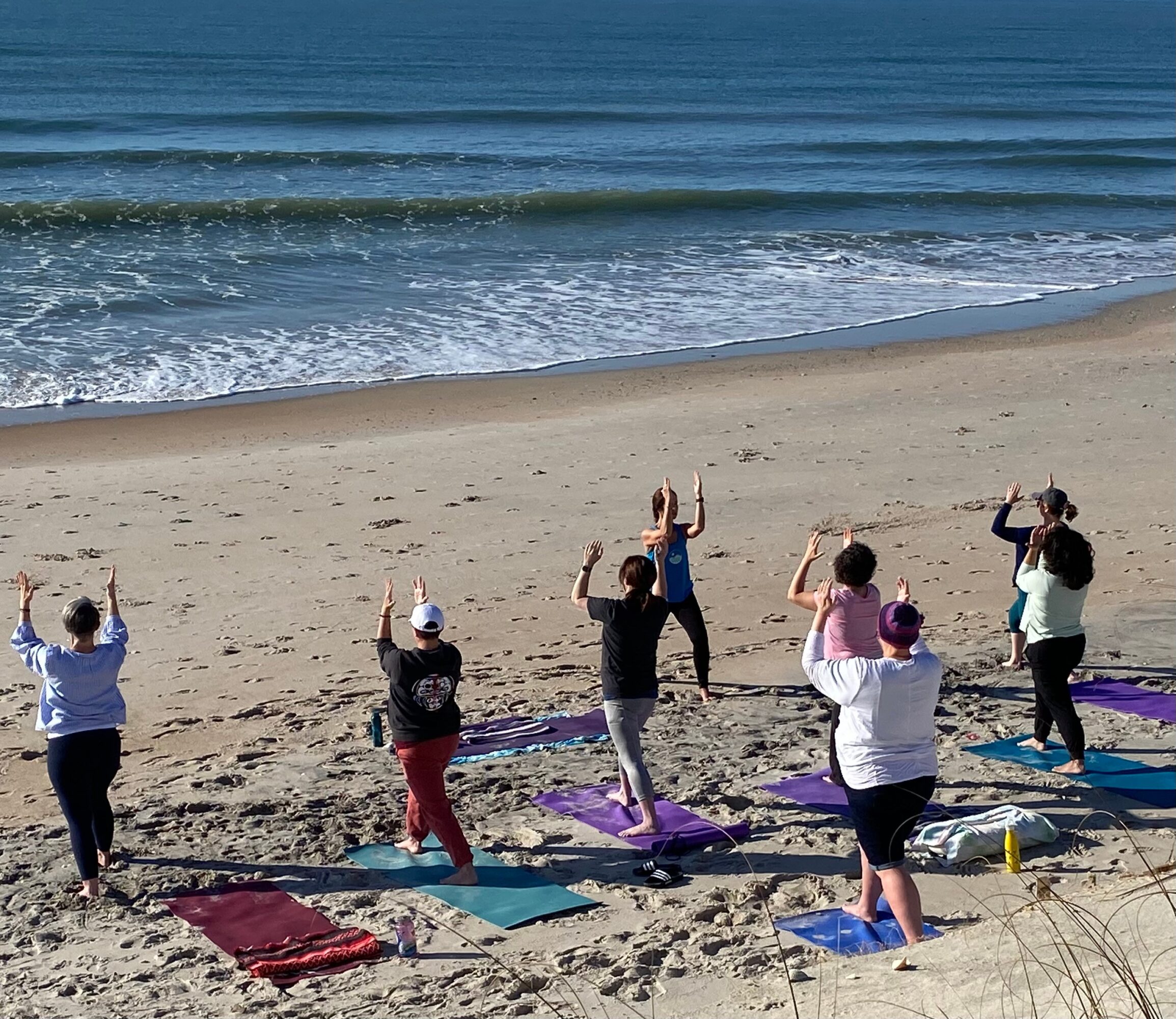 Group of people practicing yoga on the beach with ocean waves in the background, some standing and stretching, others on mats.