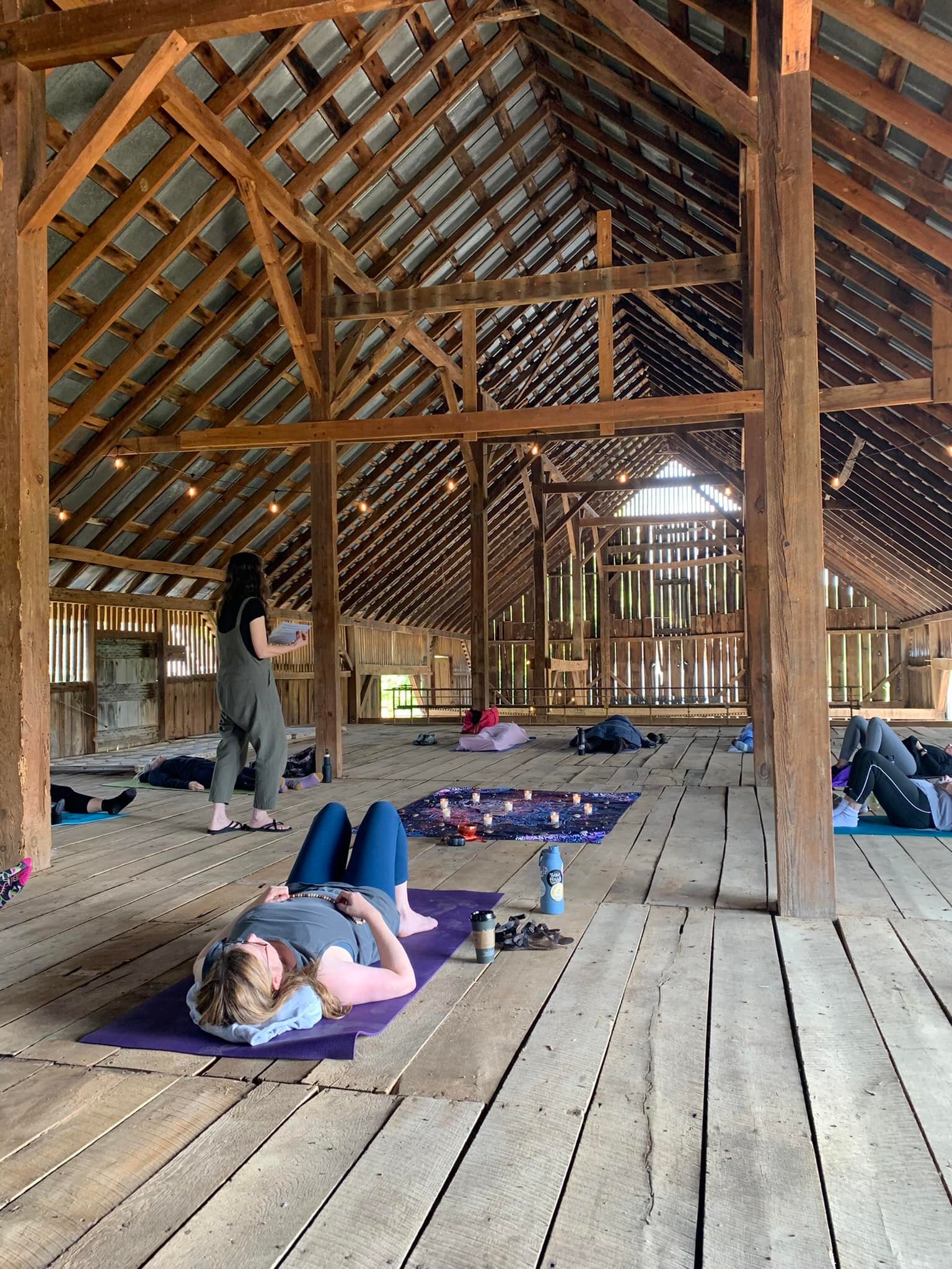People lying on mats inside a wooden structure with a high, sloped roof and open sides, some resting and some sitting.