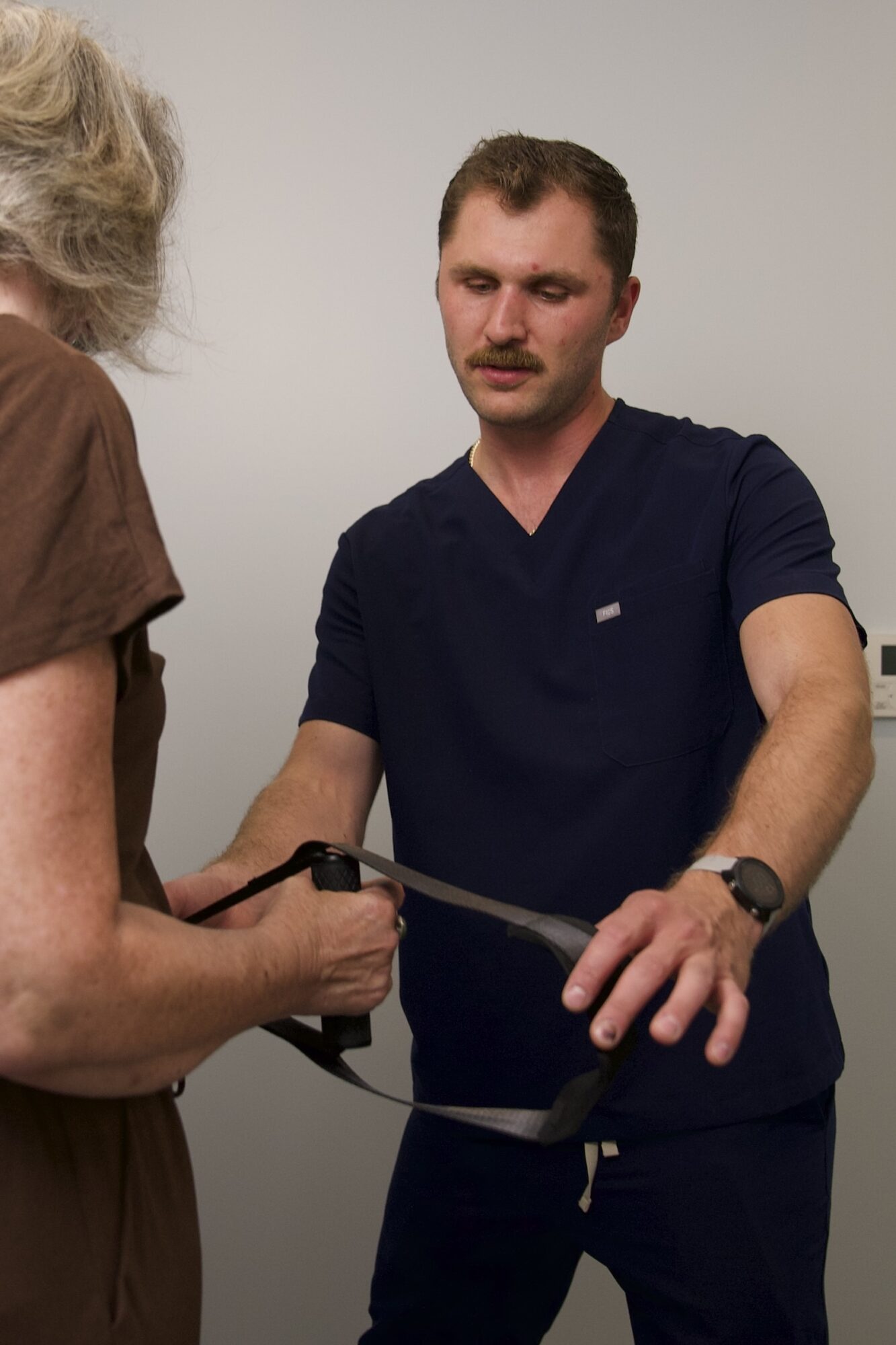 Young man with short hair and mustache wearing a navy shirt, holding a medical instrument, talking to an older woman.