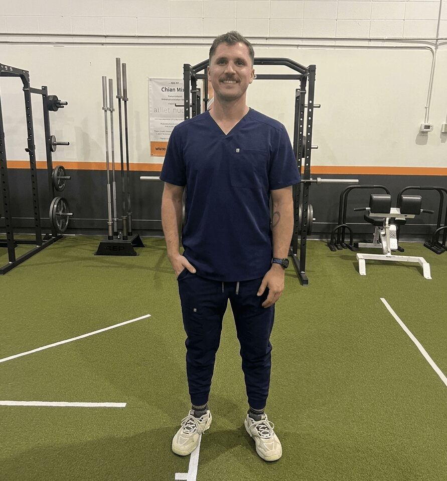 Man in navy scrubs standing in gym with equipment, smiling, hands in pockets, green floor, white walls.