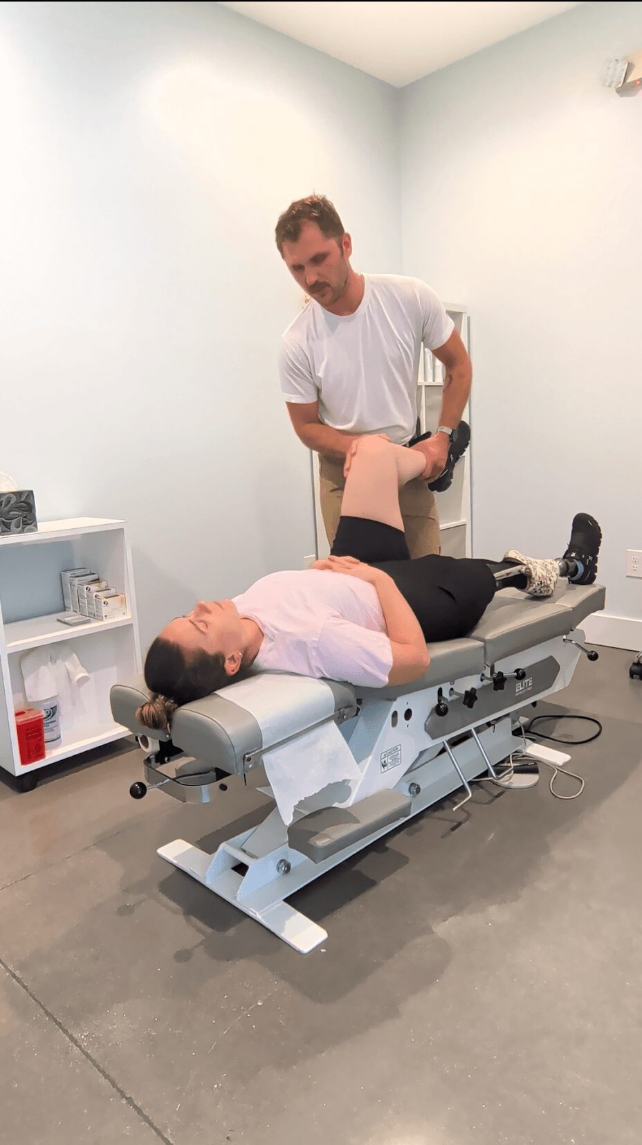 A person lying on a medical examination table with legs raised, while a healthcare professional stands beside them in a clinical room.