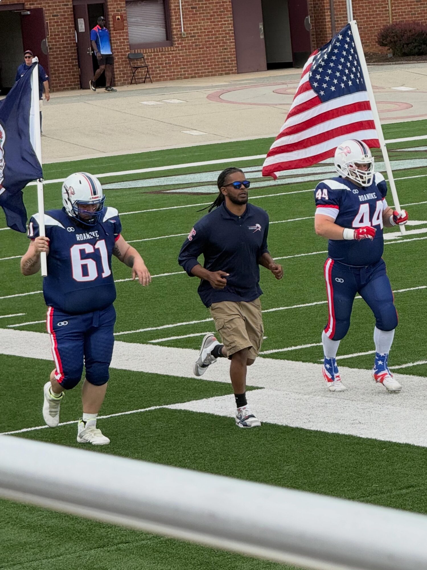 Two football players in blue uniforms and a man in casual clothing walk on a football field, one player holding an American flag.