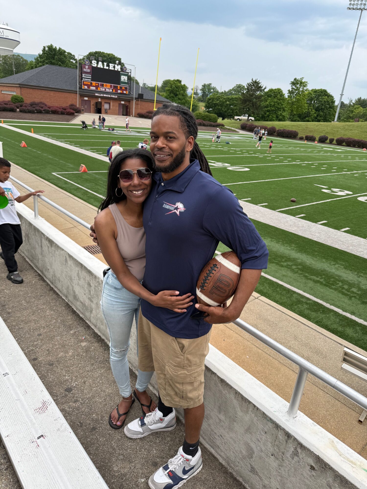 Two people standing on a sports field, one holding a football, with a building and trees in the background.