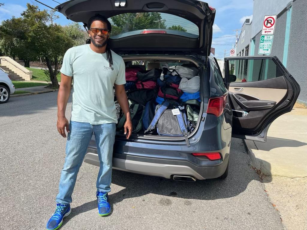 Man standing next to an open car trunk filled with luggage and bags, outdoors on a sunny day.