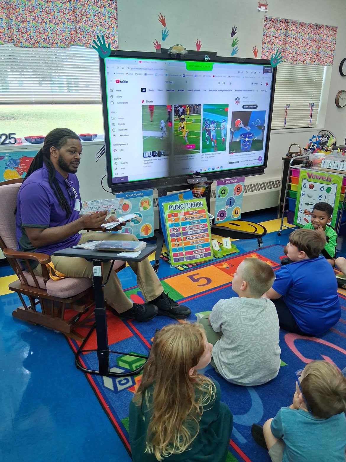 Teacher sitting in front of children on a colorful classroom rug, with a large screen displaying educational content behind him.