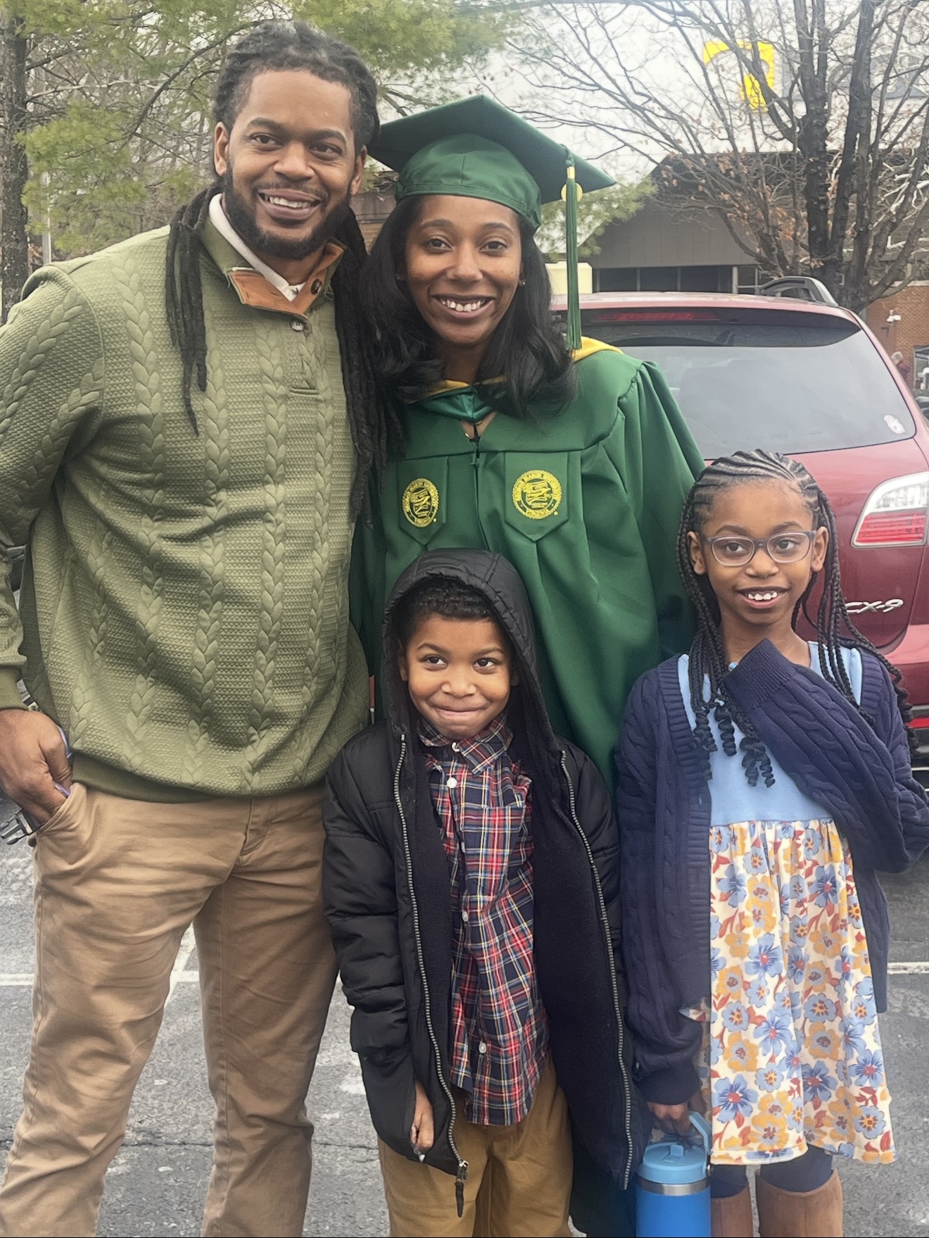 A graduate in green cap and gown standing with a man and two young girls outdoors, smiling.