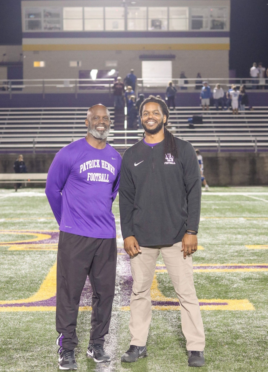 Two men standing on a football field at night, smiling, with bleachers and a building in the background.