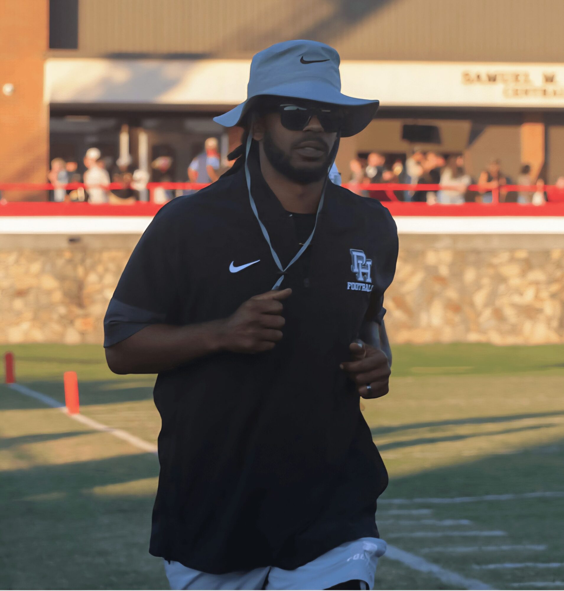 Man running on a field wearing sunglasses, a wide-brimmed hat, and a black jacket with a logo, during daytime.